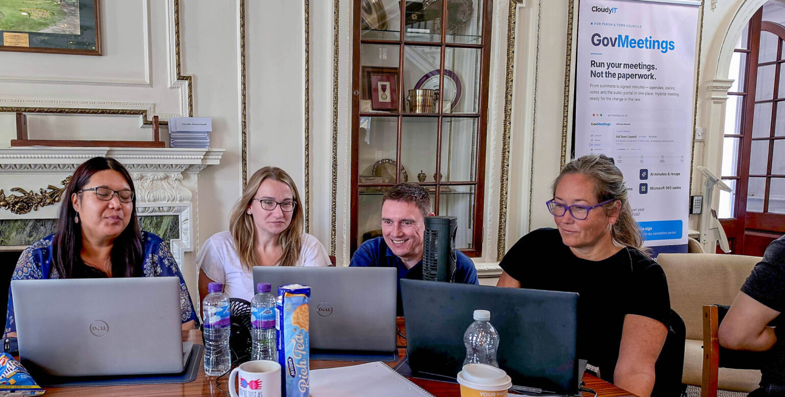 At CloudyIT's Prompt-a-thon in Dunstable, four people are seated side-by-side at a large, polished wooden conference table in an ornate room with classic decor, including a fireplace and arched display cabinet. They are looking at three open laptops, with various items like water bottles, notebooks, a mug, and a packet of biscuits scattered on the table in front of them. In the background to the right, a promotional pull-up banner for 'CloudyIT GovMeetings