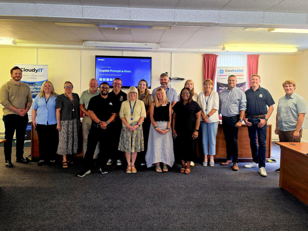 A group of sixteen CloudyIT team members and Llanelli Rural Council delegates posing together in a council meeting room for Copilot Day. They stand in front of a wall-mounted screen displaying a "Copilot Prompt-a-thon" presentation slide, flanked by CloudyIT and GovAssist branded pull-up banner stands.
