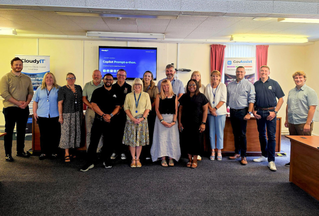 A group of sixteen CloudyIT team members and Llanelli Rural Council delegates posing together in a council meeting room for Copilot Day. They stand in front of a wall-mounted screen displaying a "Copilot Prompt-a-thon" presentation slide, flanked by CloudyIT and GovAssist branded pull-up banner stands.