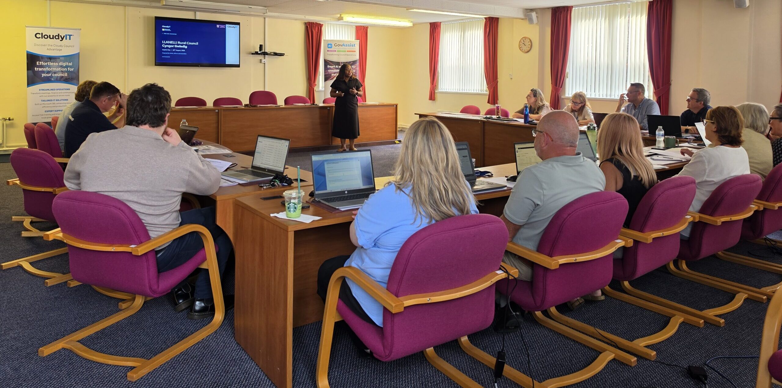 Attendees seated around wooden conference tables with laptops open, listening to a speaker presenting at the front of a meeting room between CloudyIT and GovAssist banner stands.