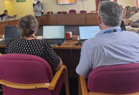 Rear view of attendees sitting in magenta office chairs at wooden desks with laptops open, facing toward the front of a meeting room. A speaker stands next to a CloudyIT banner beside a wall-mounted monitor, presenting to the seated group.